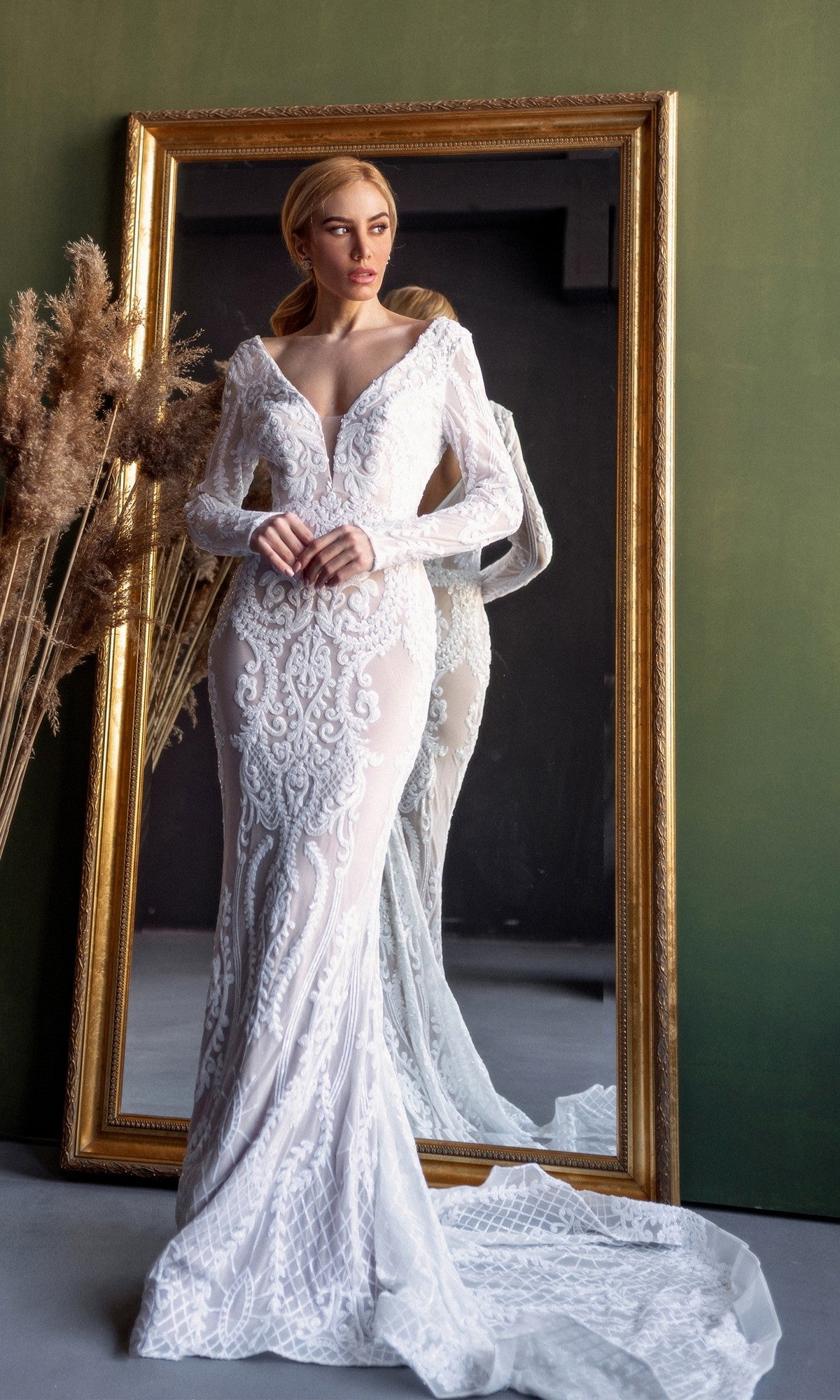 A woman wearing the Tina Holly BB021 Long Sleeve White Bridal Gown with intricate lace stands before a large gold-framed mirror, her full figure reflected.